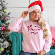 Woman wearing a pink sweatshirt with 'Season of Hope' text, sitting next to a decorated Christmas tree.
