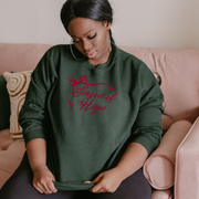 Woman wearing a green sweatshirt that says Season of Hope in burgandy with pink text sitting on a couch.