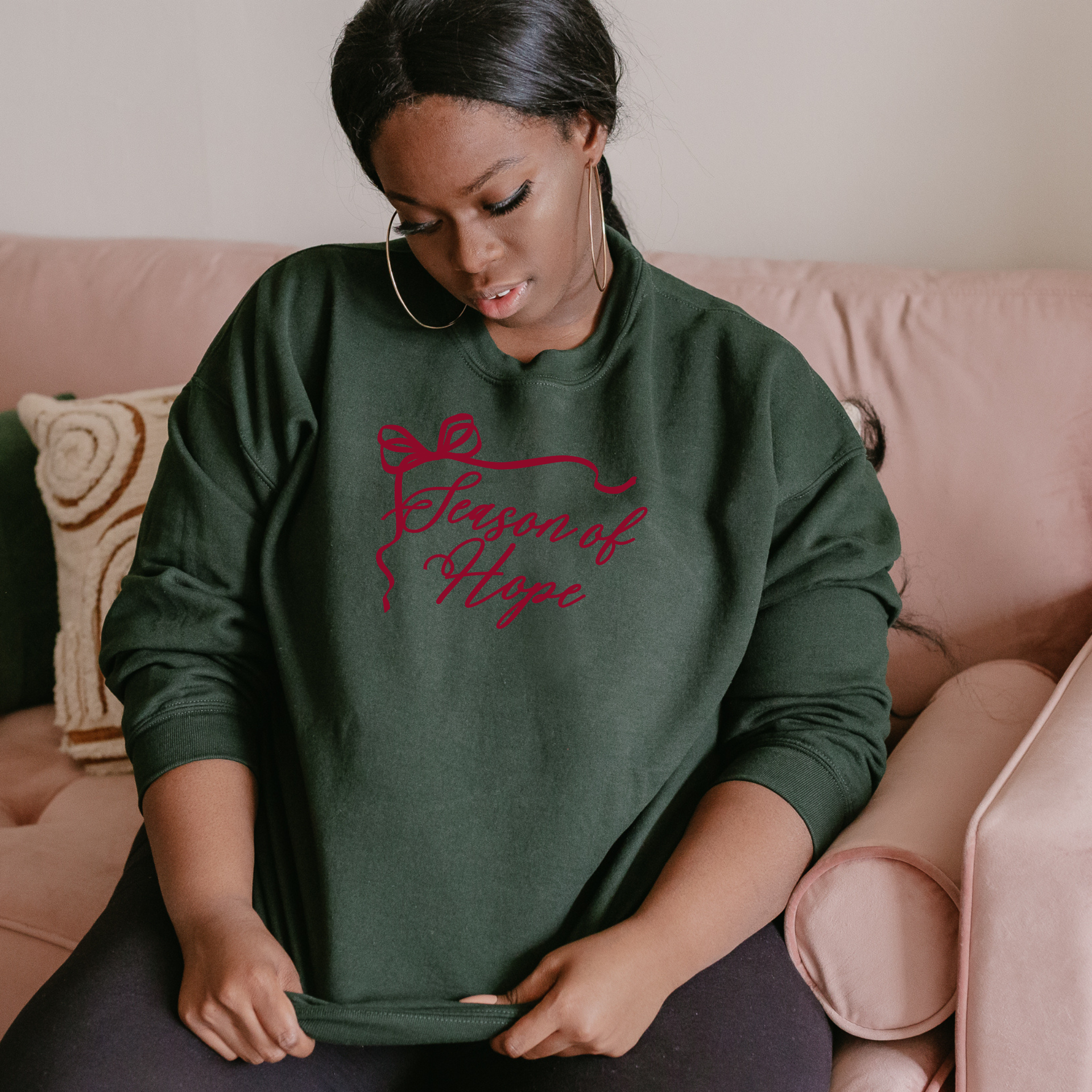 Woman wearing a green sweatshirt that says Season of Hope in burgandy with pink text sitting on a couch.