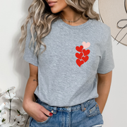 Woman wearing a light heather gray t-shirt featuring a small stacked h1 in 6 heart design in red and pink on the left chest.