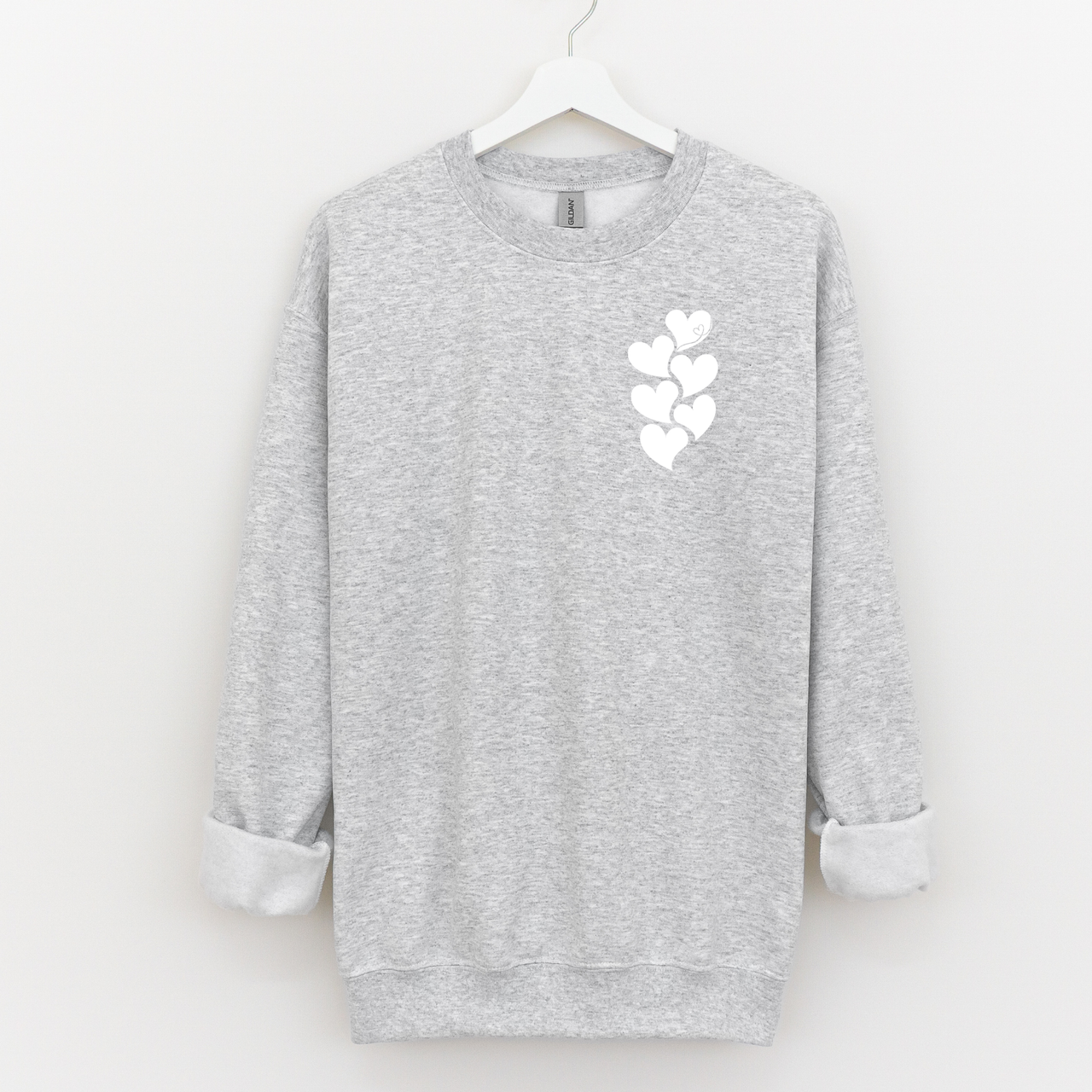 Woman wearing a beige crewneck sweatshirt with a white stacked heart “1 in 6” infertility awareness design, sitting in a cozy neutral room.
