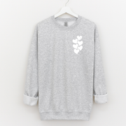 Woman wearing a beige crewneck sweatshirt with a white stacked heart “1 in 6” infertility awareness design, sitting in a cozy neutral room.