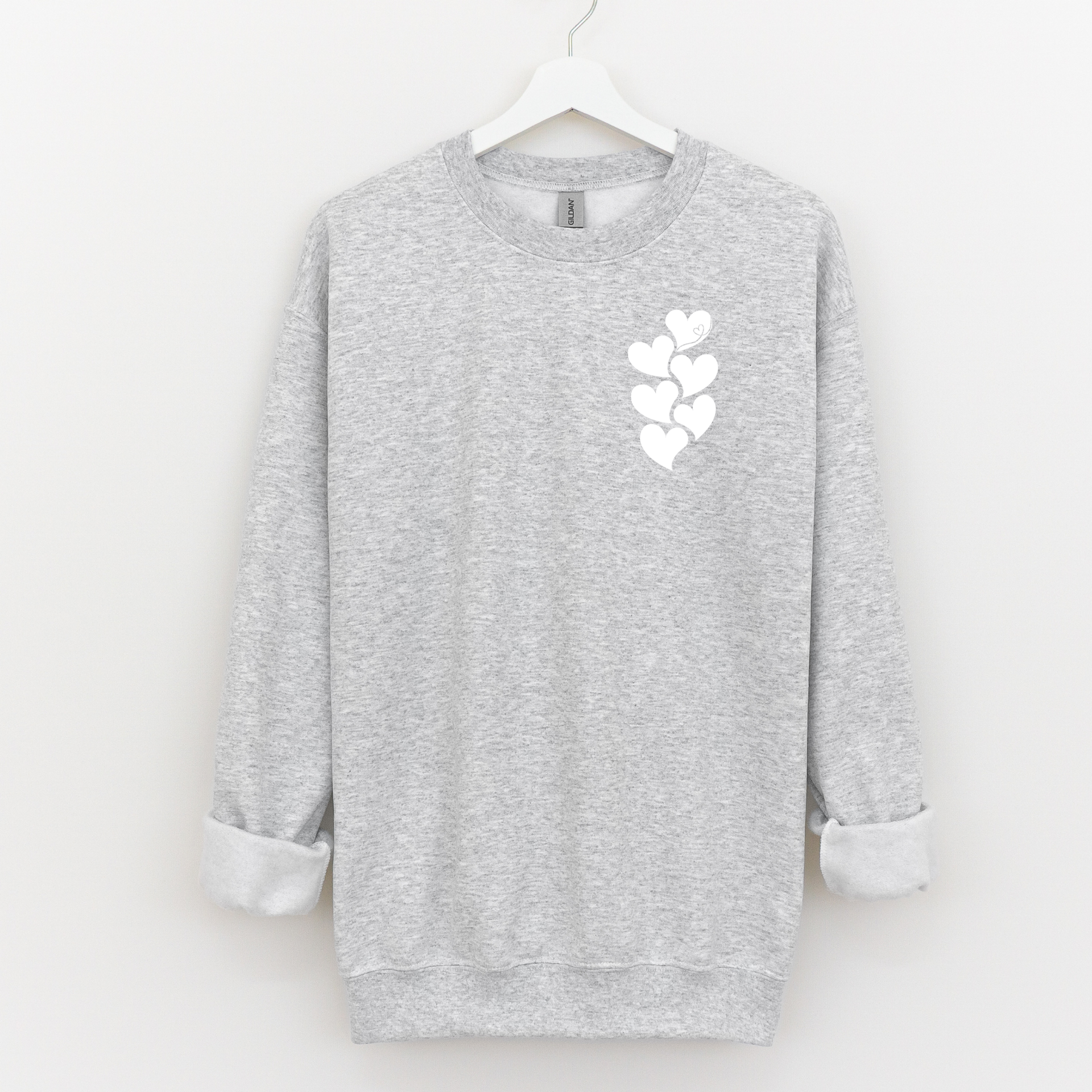 Woman wearing a beige crewneck sweatshirt with a white stacked heart “1 in 6” infertility awareness design, sitting in a cozy neutral room.