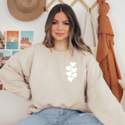 Woman wearing a beige crewneck sweatshirt with a white stacked heart “1 in 6” infertility awareness design, sitting in a cozy neutral room.