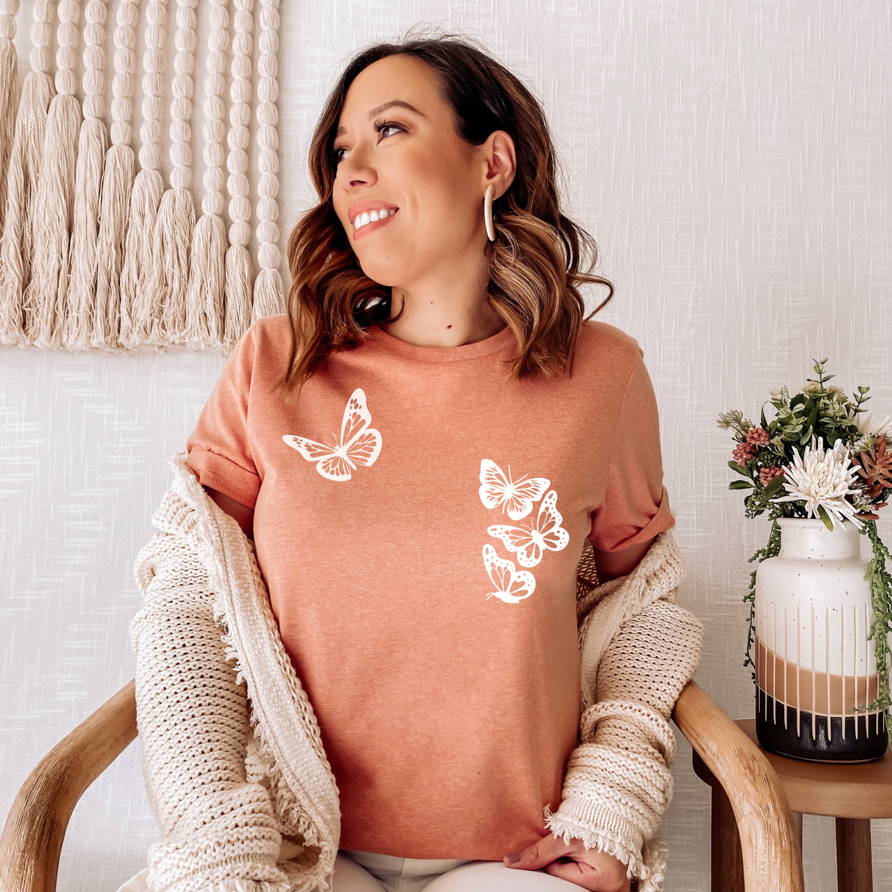 Woman wearing a sunset orange t-shirt with butterfly designs, sitting on a wooden chair. with four white butterflies symbolizing the 1 in 4 miscarriage statistic.