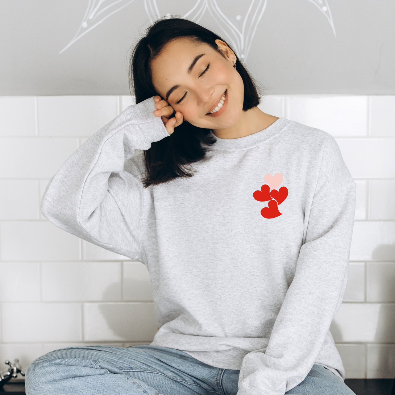Woman wearing a gray sweatshirt with a 1 in 4 cascading heart print design, sitting against a white tiled wall.