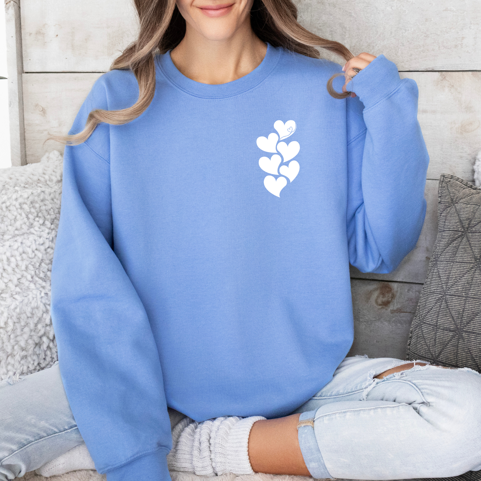 Woman wearing a beige crewneck sweatshirt with a white stacked heart “1 in 6” infertility awareness design, sitting in a cozy neutral room.