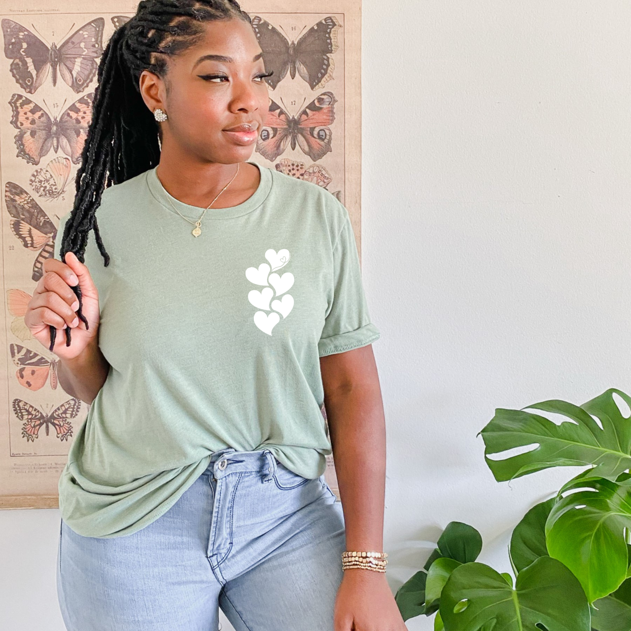 Woman wearing a sage green tee with a white stacked heart “1 in 6” infertility awareness design, sitting in a cozy neutral room.