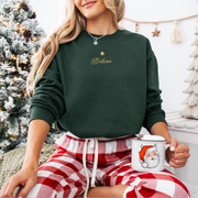 Person wearing a green sweatshirt with 'Believe' text, red plaid shorts, and holding a Santa mug in a festive setting.