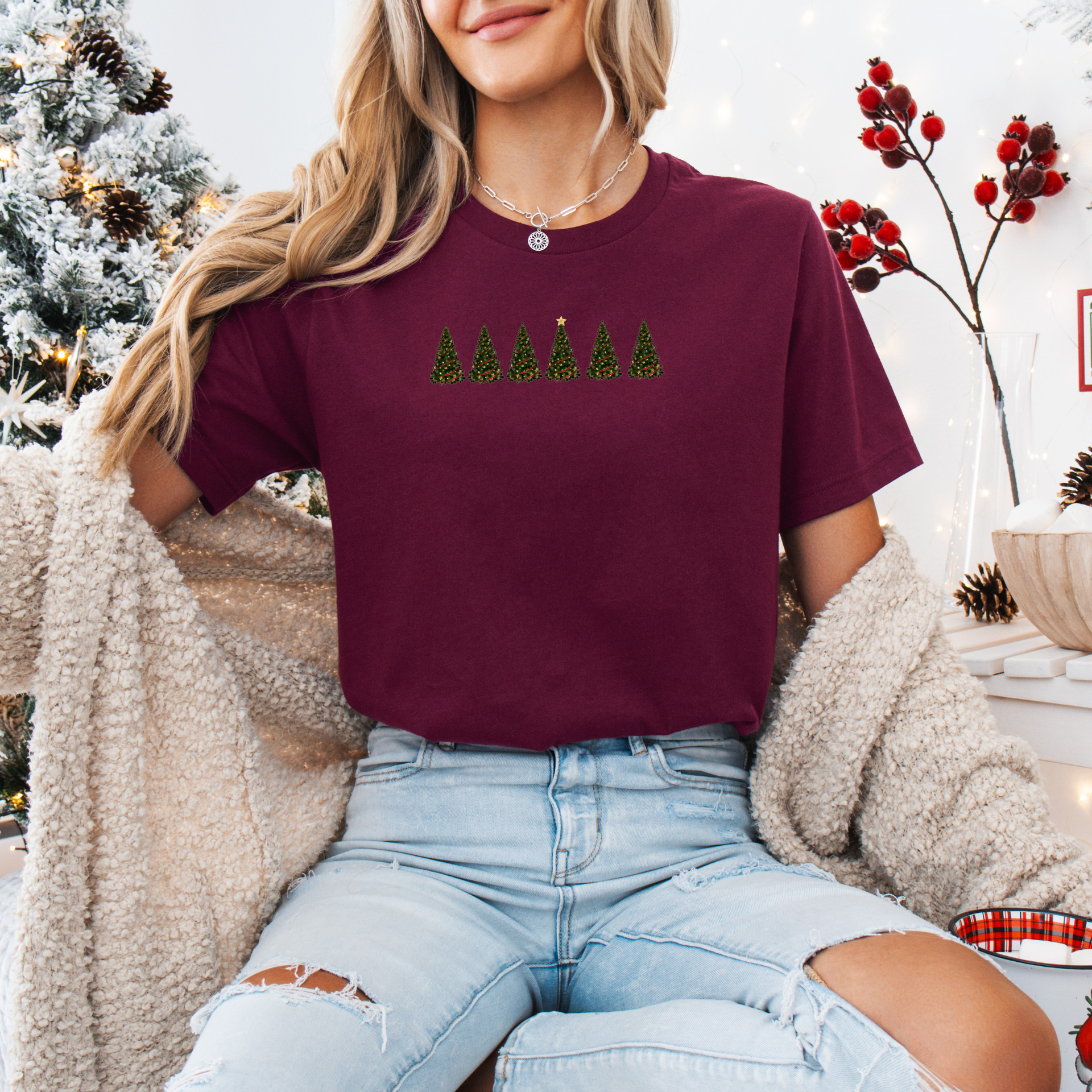 Woman wearing an maroon holiday t-shirt with a subtle 1 in 6 infertility awareness Christmas tree design, standing in a festive room with a decorated tree and wrapped gifts.