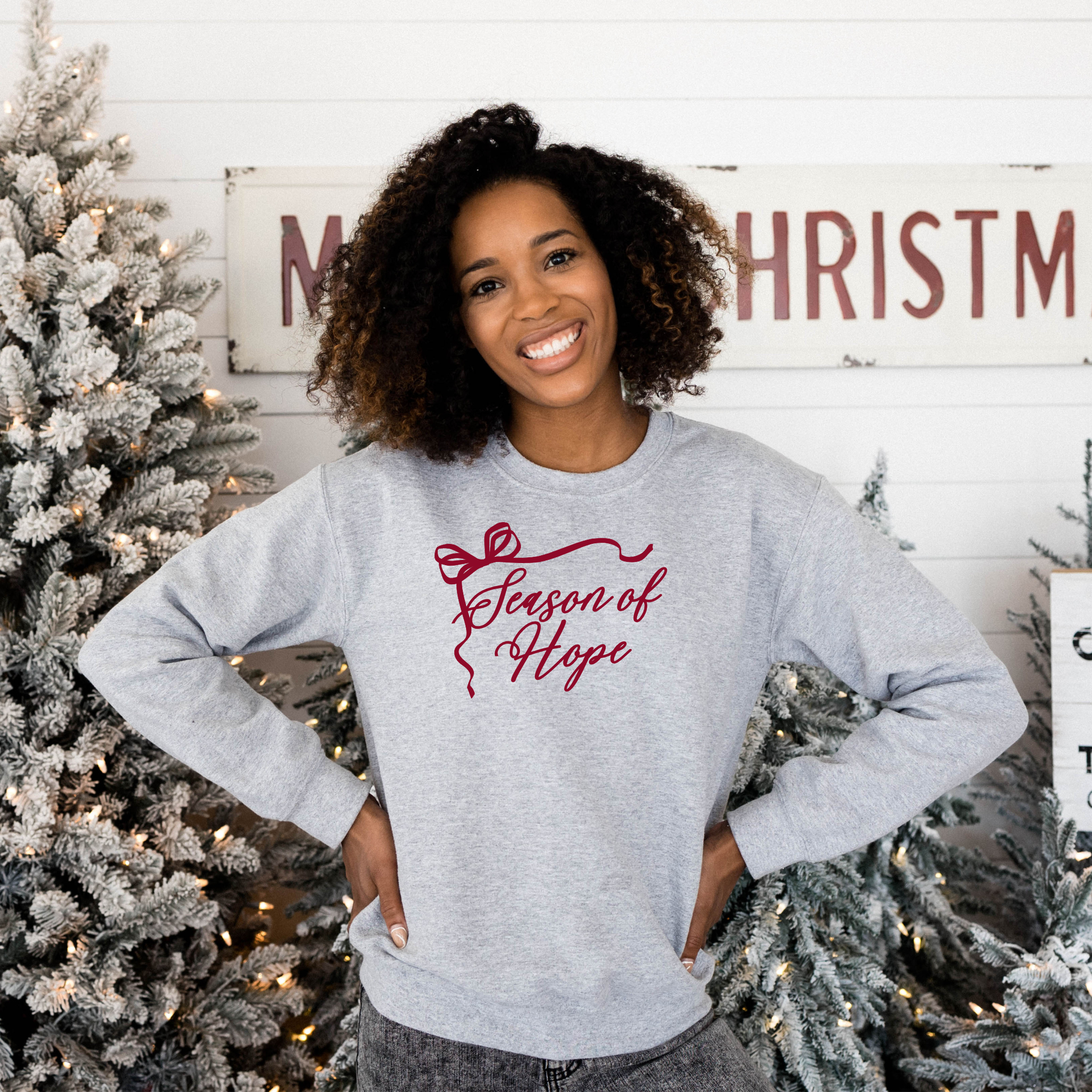 Woman wearing a gray sweatshirt with 'Season of Hope' text, standing in front of Christmas trees.