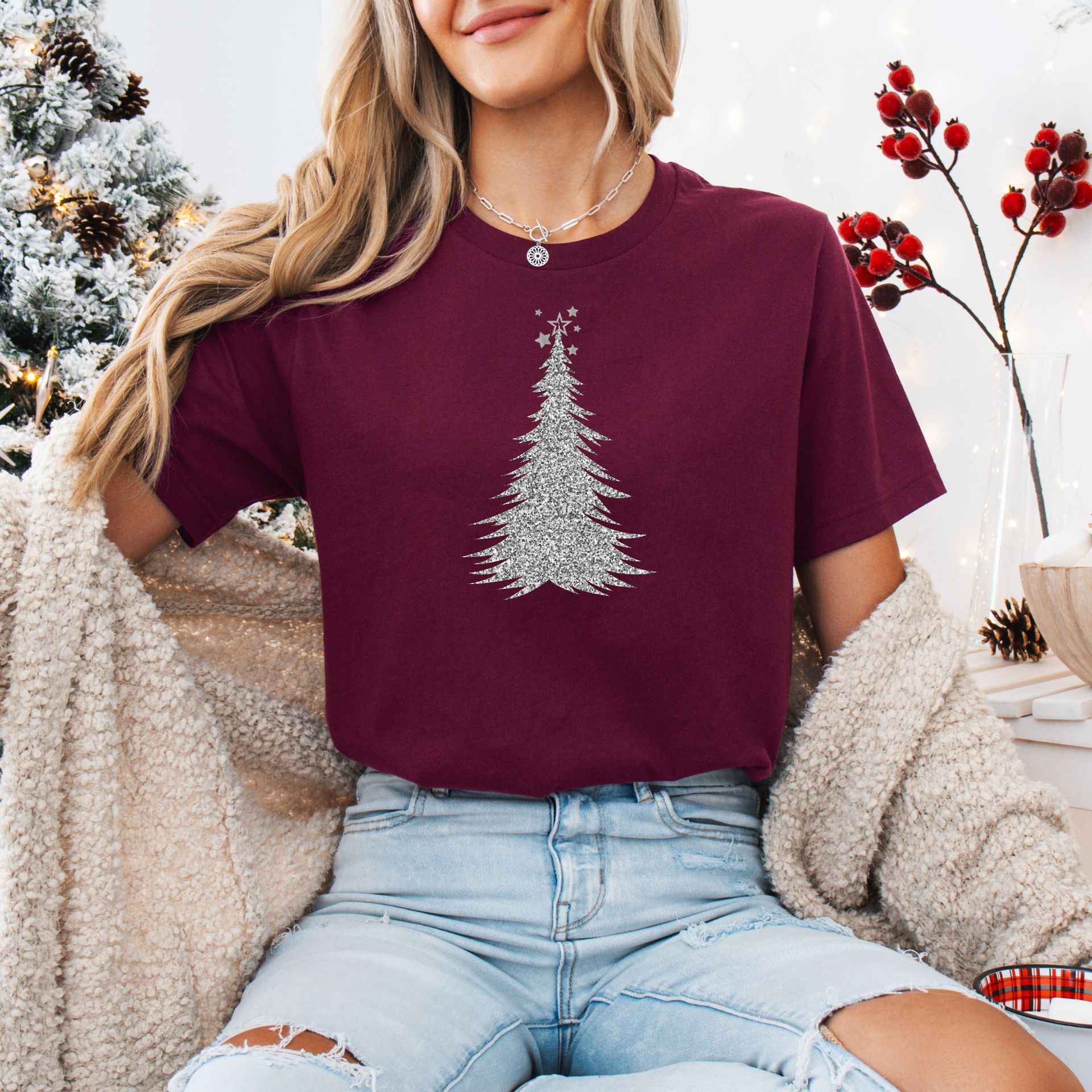 Person wearing a burgundy t-shirt with a Christmas tree design, sitting in front of a decorated Christmas tree.
